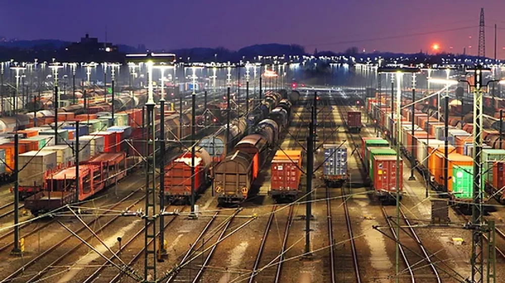 Area ferroviaria con numerosi binari, scambi e linee elettriche sullo sfondo dello skyline cittadino.