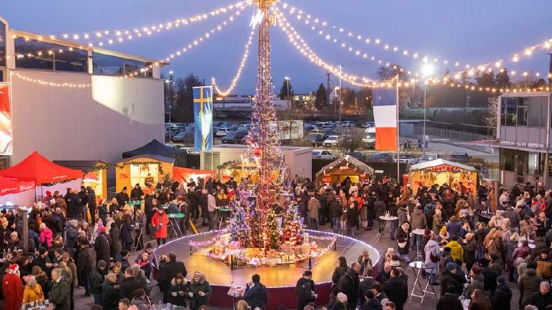 Una scena festosa da mercatino di Natale mostra un alto albero illuminato, adornato con luci e decorazioni. Una folla di persone in abiti invernali si raduna attorno alle bancarelle festive, godendosi l'atmosfera vivace e allegra. Filari di luci pendono d