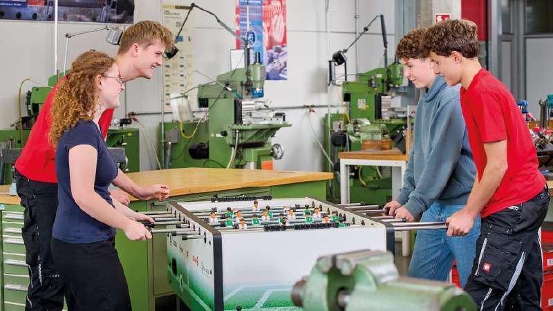 Quattro giovani, due per parte, giocano a biliardino in un capannone industriale. Sono sorridenti e concentrati, con macchinari verdi sullo sfondo.